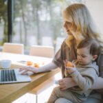 A mother works on a laptop at home while holding her baby, showcasing remote working and parenting.