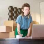 A young woman working on her laptop surrounded by cardboard boxes, indicating online business operations.